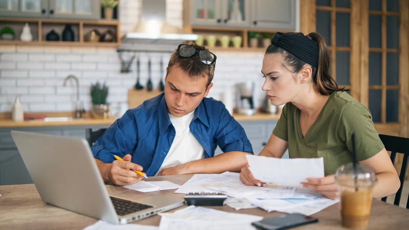 Uma mão humana segura numa caneta sobre um documento pousado numa mesa enquanto em primeiro plano está uma pequena casa